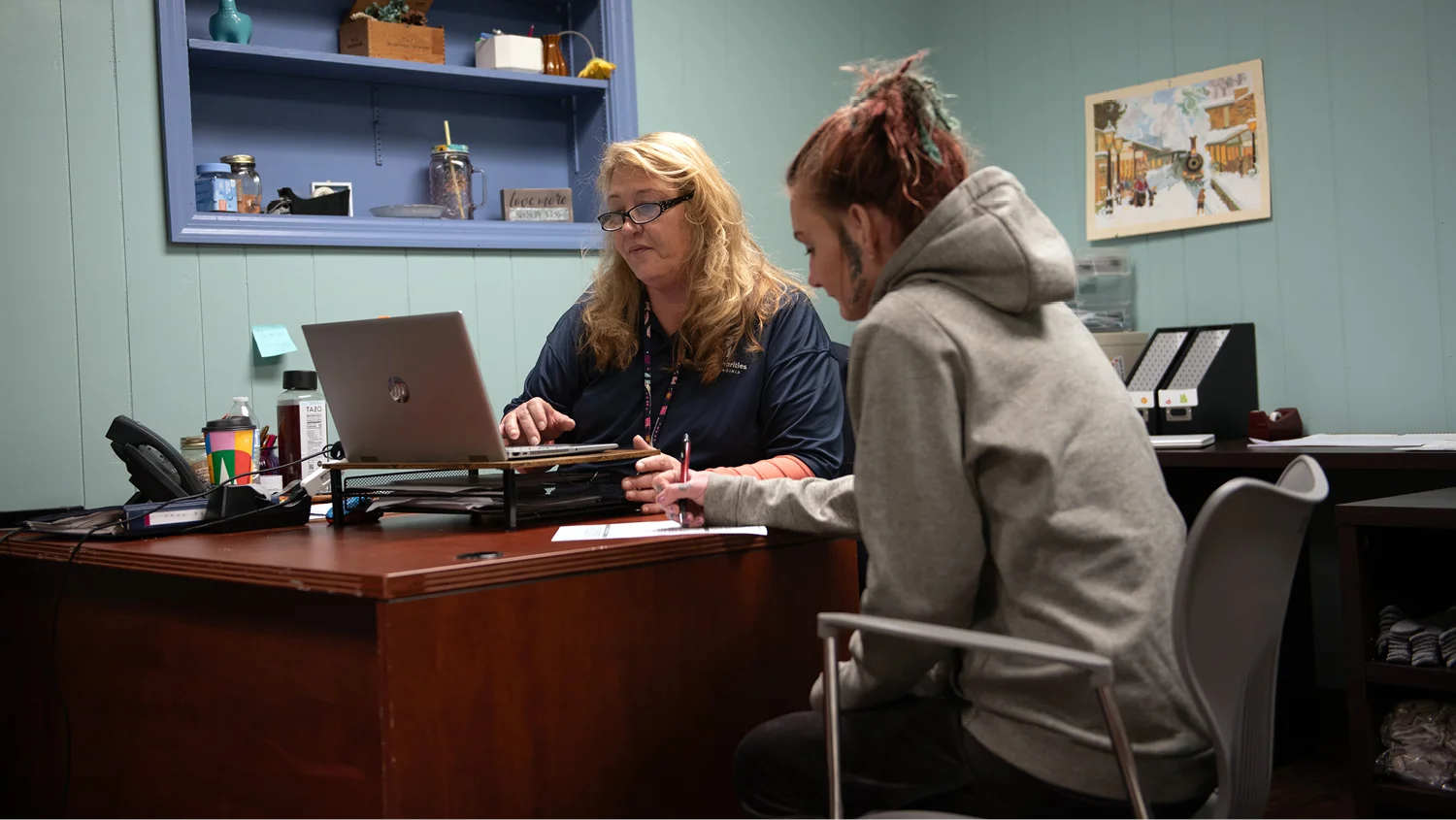 At a Catholic Charities housing site, a man with a beard in a fleece vest bends over a washing machine, checking on his clothes. Another man, wearing a purple hoodie, sits nearby.